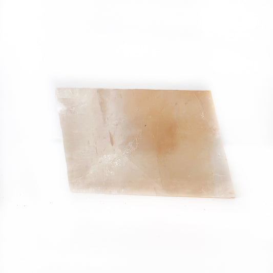 A rectangular piece of Optical Orange Calcite with translucent, warm peachy-orange tones, resting diagonally on a white surface. The smooth, polished stone displays natural lines and cloud-like inclusions, creating a soft, ethereal appearance. | Simply Esoteric
