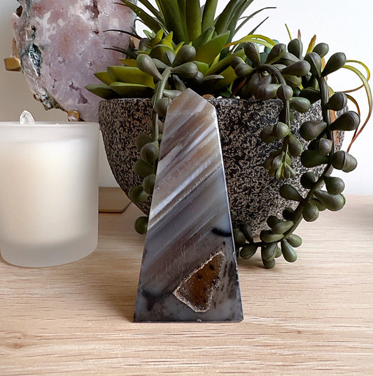 Brown agate obelisk crystal standing upright on a wooden surface, displaying layered natural brown and grey banding with a geode-like pocket at the base. | Simply Esoteric