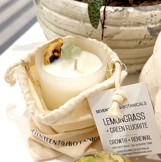 A Lemongrass + Green Fluorite crystal infused candle by Seventeen70 Botanicals. The candle is set in a frosted glass jar, adorned with a piece of Green Fluorite crystal and dried lemongrass on top. It is placed in a cream-colored drawstring bag with the brand's name printed on it. The candle's label indicates it is vegan-friendly, made of soy wax, and has an 18+ hour burn time. The background includes a potted plant and additional crystal decor. | Simply Esoteric