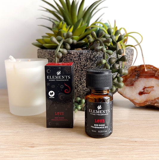 A 10ml bottle of Elements Love Fragrance Oil with a red rose scent, shown next to its matching red and black box packaging featuring celestial illustrations. Displayed on a wooden surface with a white candle, succulent plant, and polished agate slice, creating a romantic and serene setting. | Simply Esoteric
