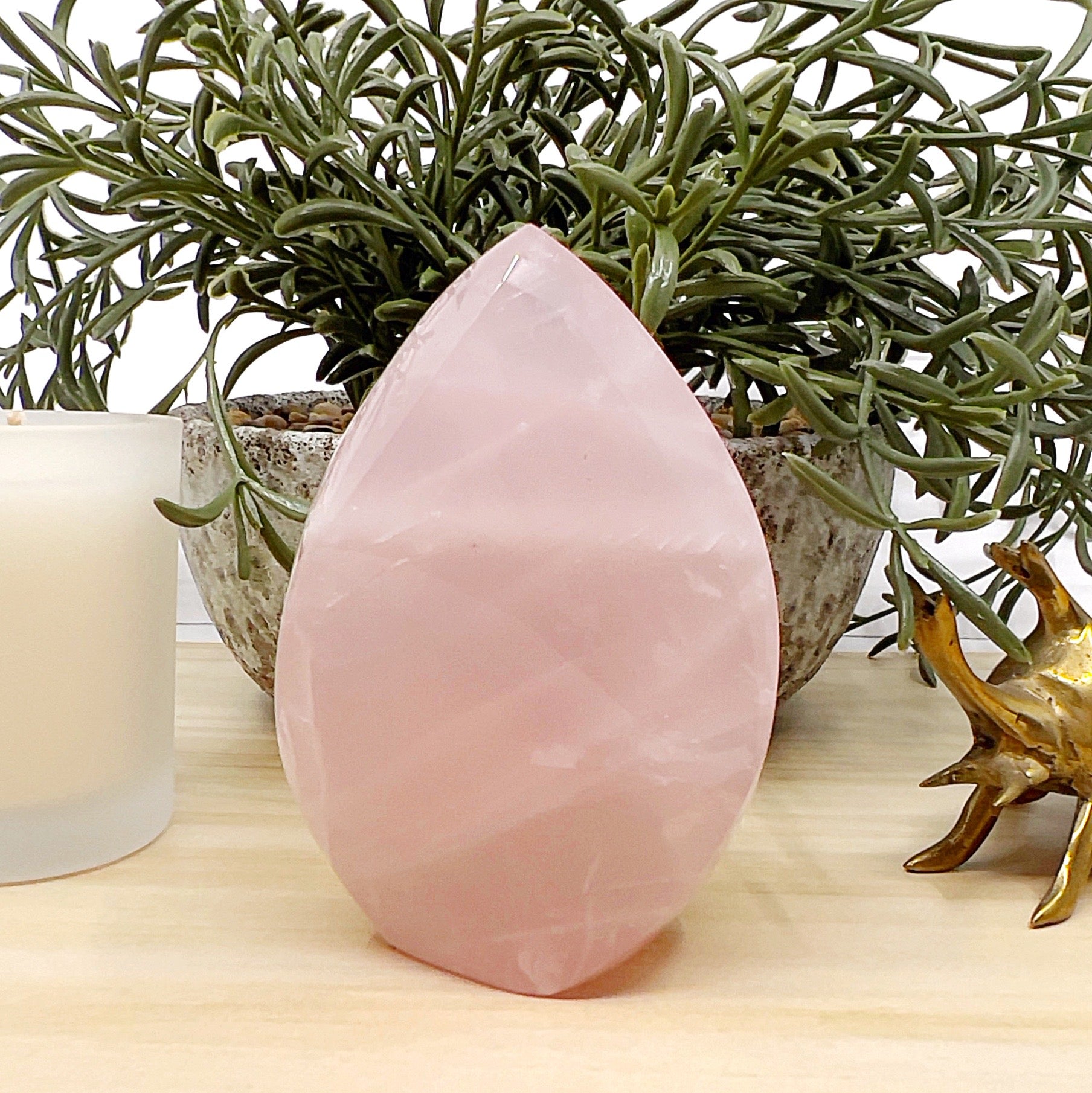 Polished 9cm rose quartz flame with soft pink tones and natural variations, displayed on a light wooden surface with a white candle, ceramic pot with greenery, and a small brass beetle ornament in the background. | Simply Esoteric