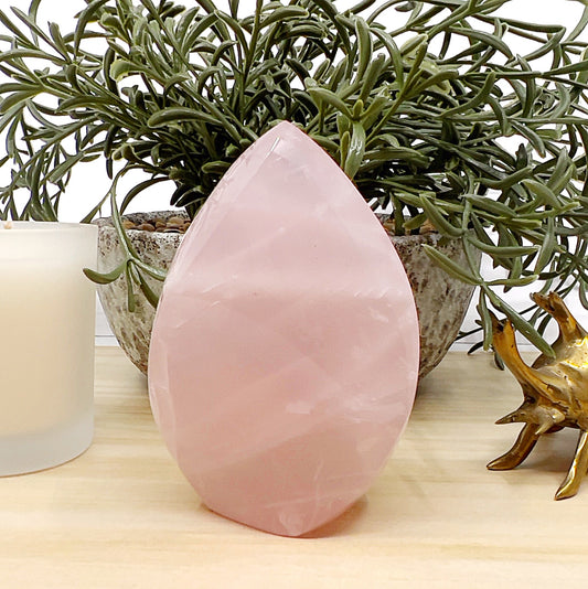 Polished 9cm rose quartz flame with soft pink tones and natural variations, displayed on a light wooden surface with a white candle, ceramic pot with greenery, and a small brass beetle ornament in the background. | Simply Esoteric