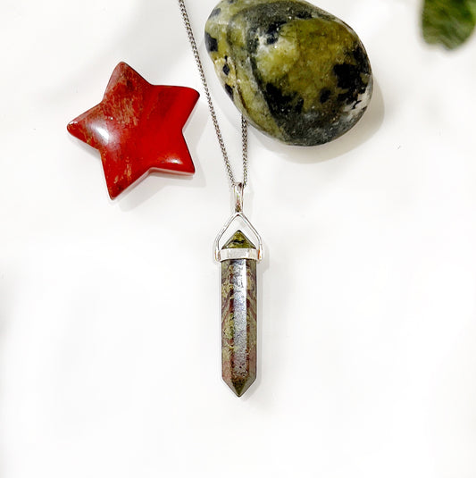 A close-up image of a Dragon's Blood Jasper and sterling silver double terminated point pendant. The pendant features a vibrant green and red jasper crystal set in a sleek sterling silver frame, with pointed ends that enhance the stone's energy. It is displayed alongside a red jasper star and a green jasper stone, highlighting the natural beauty of the crystals. The pendant does not include a chain. | Simply Esoteric
