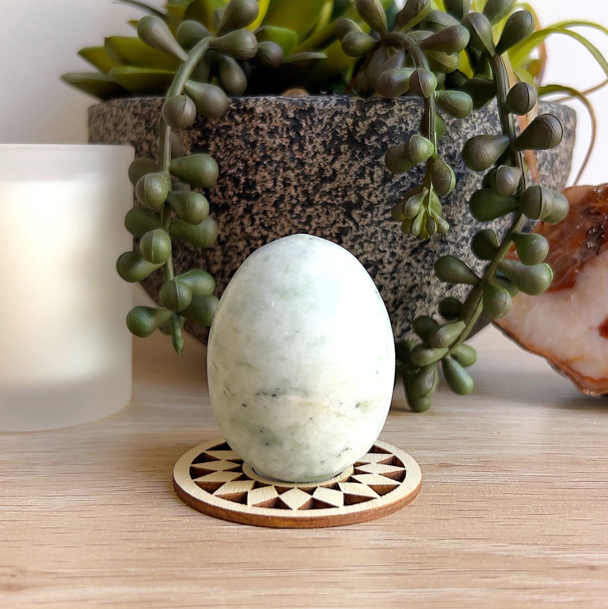 A polished jadeite egg approximately 5cm tall, featuring soft pale green tones with gentle white marbling. Displayed upright on a wooden geometric base, set against a natural backdrop with succulents, a candle, and agate slice for a calming, earthy presentation. | Simply Esoteric