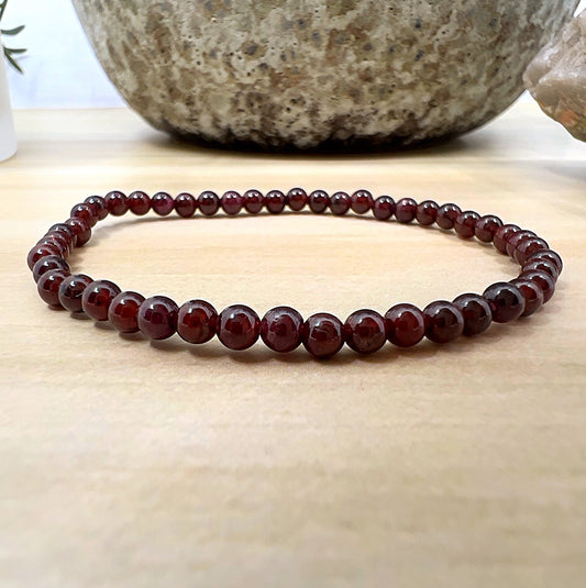 Handmade Garnet crystal bead bracelet with 4mm round beads on stretchy cord, featuring deep red polished stones, displayed on a wooden surface. | Simply Esoteric