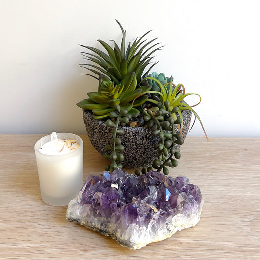 A vibrant purple amethyst crystal cluster, approximately 14cm long, displayed next to a lush succulent arrangement and a white candle, creating a serene and calming atmosphere. | Simply Esoteric