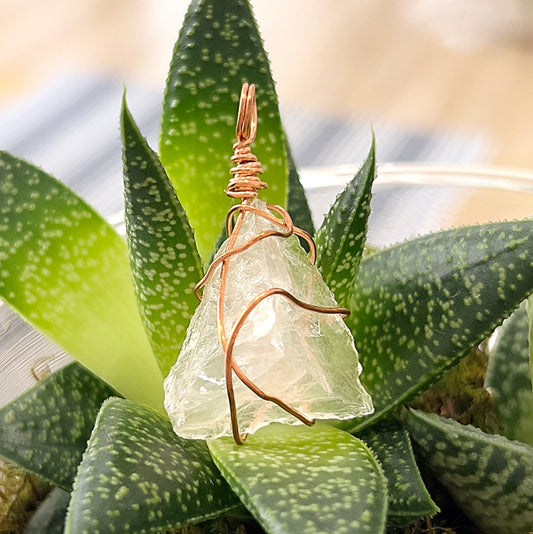 A close-up image of a rose quartz wire wrap pendant in rose gold colour, set against a background of green succulents. The soft pink rose quartz crystal is encased in delicate rose gold-coloured wire, showcasing its natural beauty and elegance. | Simply Esoteric
