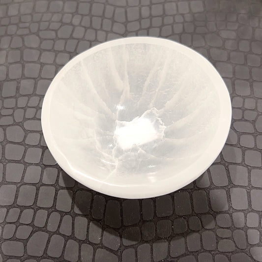 A small, circular selenite bowl resting on a dark, hexagonal-patterned surface. The bowl is translucent with a milky white appearance, showcasing natural striations and a smooth finish that captures the light, creating a serene and inviting glow. | Simply Esoteric