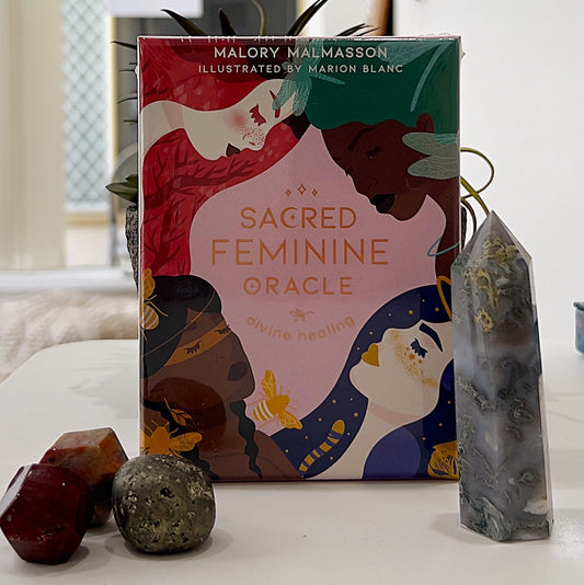A box of the *Sacred Feminine Oracle* by Malory Malmasson, illustrated by Marion Blanc, displayed on a white surface with decorative gemstones and a crystal tower nearby. The box features vibrant illustrations of diverse feminine figures with a harmonious pink and earthy-toned design, highlighting themes of divine healing and empowerment. The setup is accompanied by natural and mystical decor elements, emphasizing spirituality and connection. | Simply Esoteric