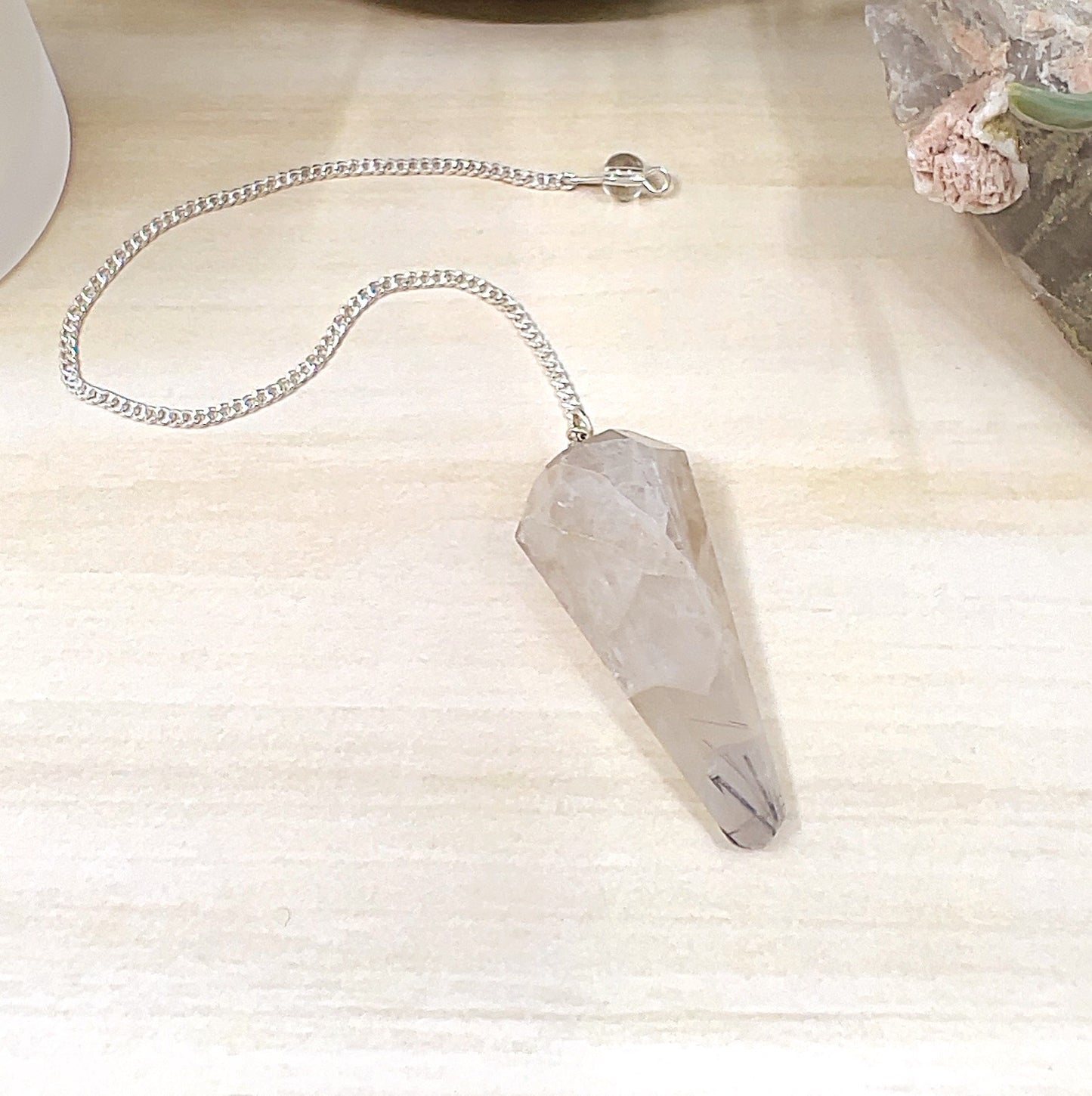 Faceted tourmalinated quartz pendulum with clear quartz and natural black tourmaline inclusions, attached to a metal link chain with bead detail, displayed on a light wooden surface with a white candle, crystal cluster, and ceramic pot in the background. | Simply Esoteric