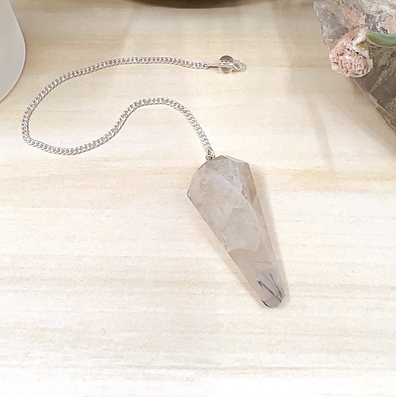Faceted tourmalinated quartz pendulum with clear quartz and natural black tourmaline inclusions, attached to a metal link chain with bead detail, displayed on a light wooden surface with a white candle, crystal cluster, and ceramic pot in the background. | Simply Esoteric
