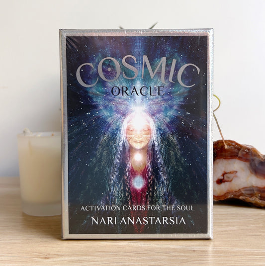 A boxed set of “Cosmic Oracle: Activation Cards for the Soul” by Nari Anastarsia, featuring ethereal artwork of a radiant cosmic being with glowing chakras. Displayed on a wooden surface with a candle, plant, and agate slice in the background, creating a serene and spiritual setting. | Simply Esoteric