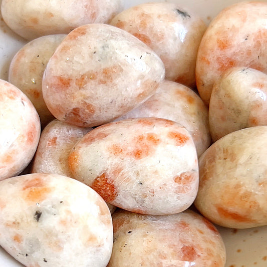 A collection of polished sunstone tumbled stones with a smooth finish and an interplay of orange and cream hues. Some stones exhibit natural sparkling inclusions and flecks of darker mineral deposits, adding to their unique charm. The stones vary slightly in shape and size, with a warm and inviting appearance reminiscent of a sunlit day. | Simply Esoteric