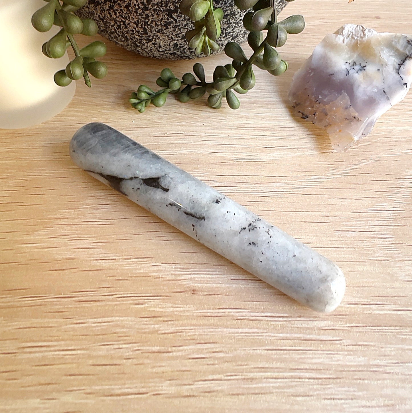 A polished moonstone massage wand measuring approximately 11cm, featuring a creamy white surface with natural black veining, displayed on a light wooden surface with a candle, plant, and raw crystal in the background. | Simply Esoteric