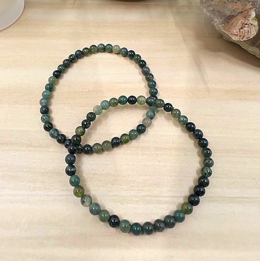 Moss Agate crystal bead bracelet with 4mm round beads on stretchy cord, featuring natural green tones with earthy inclusions, displayed on a wooden surface. | Simply Esoteric