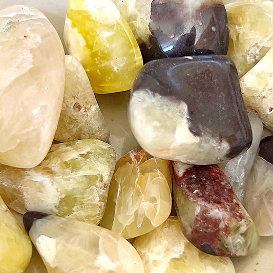 A collection of tumbled prehnite stones from Western Australia, showcasing varying shades of soft green intermixed with patches of earthy tones. The stones have a translucent, waxy luster and are randomly scattered, highlighting their natural, smooth shapes and soothing color palette. | Simply Esoteric