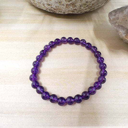 Amethyst crystal bead bracelet with polished 6mm A Grade purple beads on stretchy cord, displayed on a light wooden surface. | Simply Esoteric