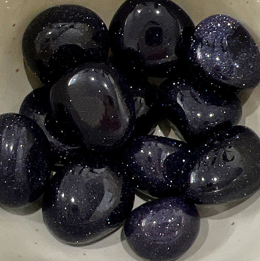 A collection of polished Blue Goldstone tumbled stones, shimmering with tiny copper flecks against a deep blue background. The stones are smooth and vary in size, with an enchanting sparkle that resembles a starry night sky. They are displayed together, showcasing their glossy finish and captivating beauty. | Simply Eosteric