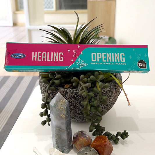 A rectangular box of Ullas Healing Premium Masala Incense, weighing 15g, displayed in a decorative setting with a natural succulent plant and a selection of crystals, including polished and pointed stones, arranged artfully in the background. The packaging is vibrant, featuring pink and teal tones with "Healing" and "Opening" prominently written. | Simply Esoteric
