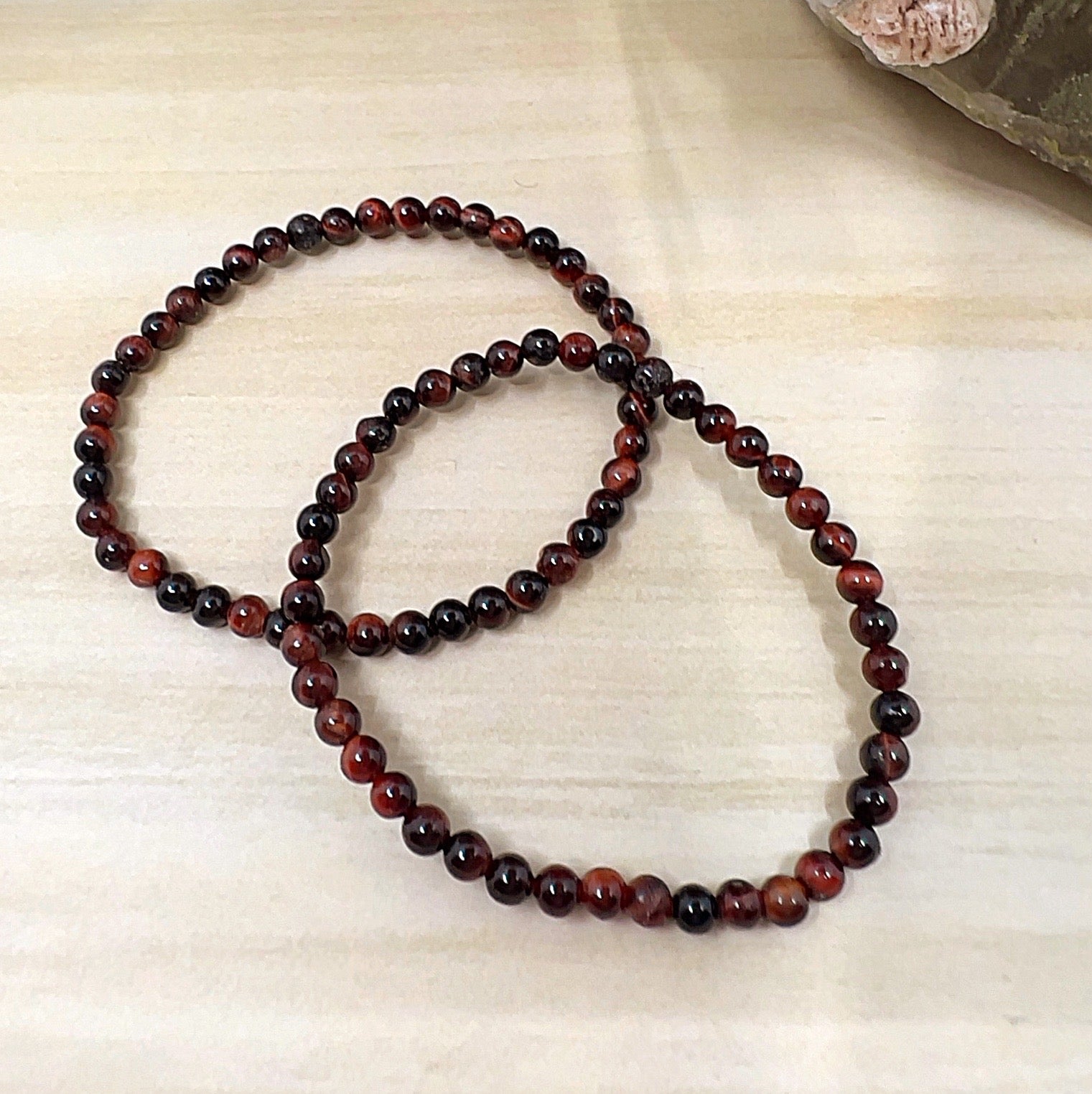 Handmade Red Tiger Eye crystal bead bracelets with 4mm round beads on stretchy cord, featuring deep reddish-brown stones with natural shimmer, displayed on a wooden surface. | Simply Esoteric