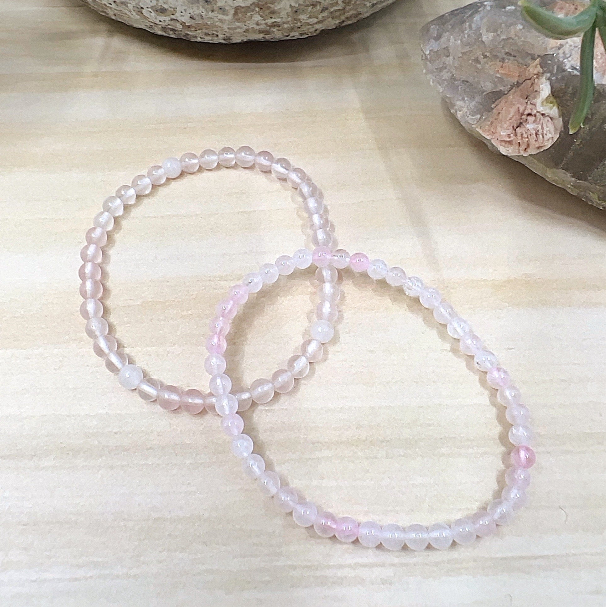 A delicate Rose Quartz crystal bead bracelet handmade with 4mm soft pink beads on a stretchy cord, displayed on a wooden surface with crystals in the background. | Simply Esoteric
