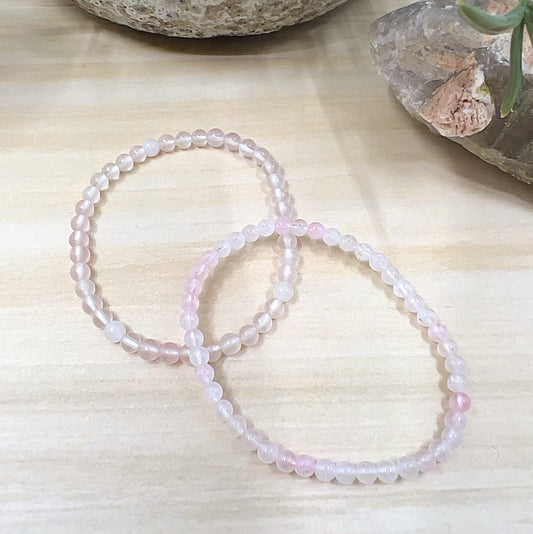 A delicate Rose Quartz crystal bead bracelet handmade with 4mm soft pink beads on a stretchy cord, displayed on a wooden surface with crystals in the background. | Simply Esoteric