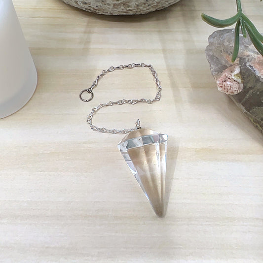 Faceted clear quartz pendulum approximately 4cm x 2cm with a metal link chain, displayed on a light wooden surface with a white candle, crystal cluster, and ceramic pot in the background. | Simply Esoteric