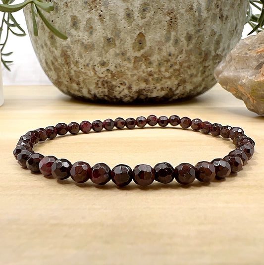 Handmade Garnet faceted crystal bead bracelet with deep red polished stones on stretchy cord, featuring sparkling light-reflective facets, displayed on a wooden surface. | Simply Esoteric