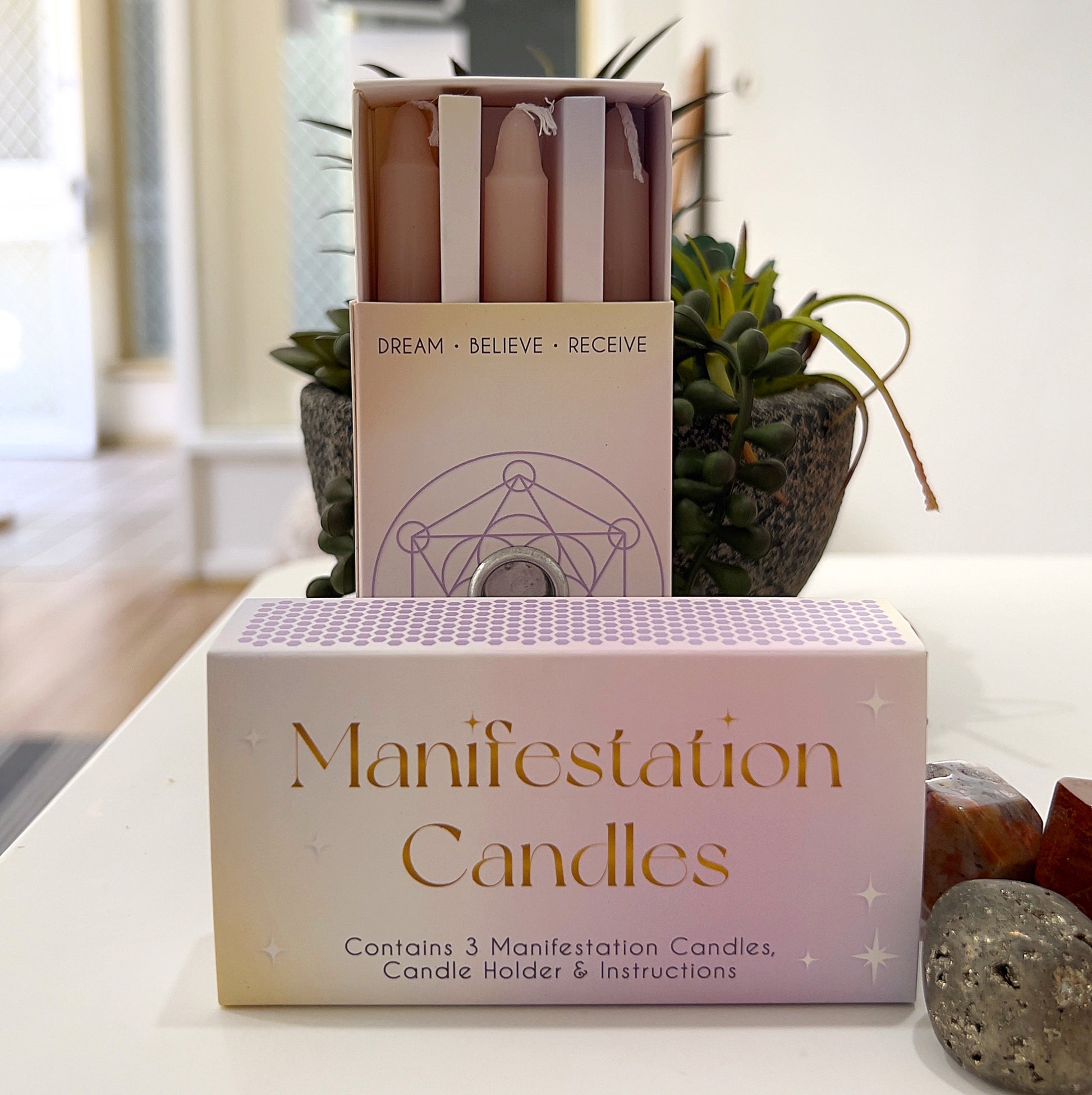 A boxed set of three pastel-colored Manifestation Candles displayed alongside their packaging. The box is labeled "Manifestation Candles" and includes a candle holder and instructions for use. The candles are designed for intention-setting and manifestation rituals, sitting elegantly on a white surface with decorative crystals and a small potted plant in the background. | Simply Esoteric