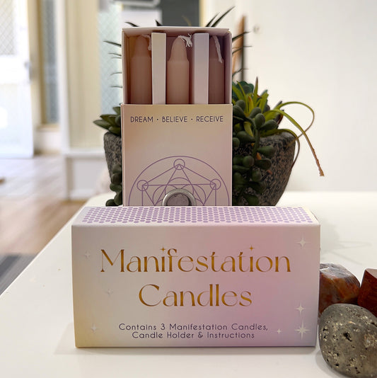 A boxed set of three pastel-colored Manifestation Candles displayed alongside their packaging. The box is labeled "Manifestation Candles" and includes a candle holder and instructions for use. The candles are designed for intention-setting and manifestation rituals, sitting elegantly on a white surface with decorative crystals and a small potted plant in the background. | Simply Esoteric