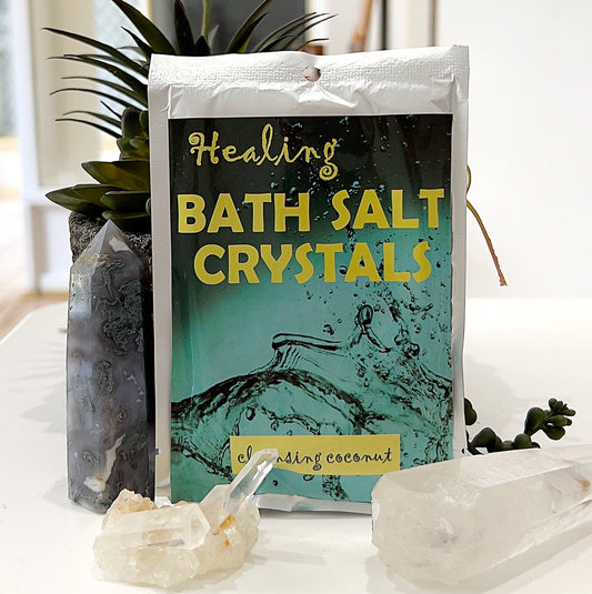 A 100g packet of Healing Bath Salt Crystals in a coconut scent, placed on a display table alongside decorative crystals and greenery. The packaging has a vibrant design with the words "Healing Bath Salt Crystals" and "cleansing coconut," evoking a refreshing and therapeutic ambiance. | Simply Esoteric