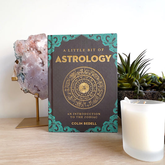 A hardcover book titled “A Little Bit of Astrology: An Introduction to the Zodiac” by Colin Bedell, featuring a gold zodiac wheel design on a black and teal cover. The book is displayed upright on a wooden surface with a white candle and crystal decor beside it. | Simply Esoteric