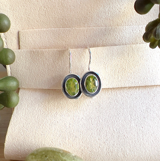 Sterling silver Peridot earrings with oval faceted green stones, displayed on a light surface with greenery accents. | Simply Esoteric