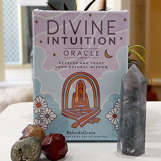 A neatly arranged box of Divine Intuition Oracle Cards by Belinda Grace, featuring a pastel-coloured cover illustration of a meditative woman surrounded by floral and celestial designs. The tagline reads, "Develop and Trust Your Natural Wisdom." The box is placed on a light surface, accompanied by crystal gemstones and a polished crystal tower, enhancing the serene and spiritual aesthetic of the image. | Simply Esoteric