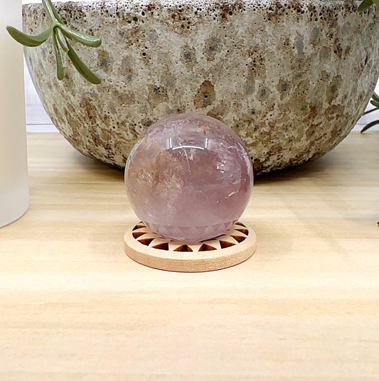 Polished amethyst and smoky quartz sphere approximately 3cm in diameter, featuring swirls of purple and earthy smoky tones, displayed on a wooden stand against a light wooden surface, with a ceramic pot and white candle in the background. | Simply Esoteric