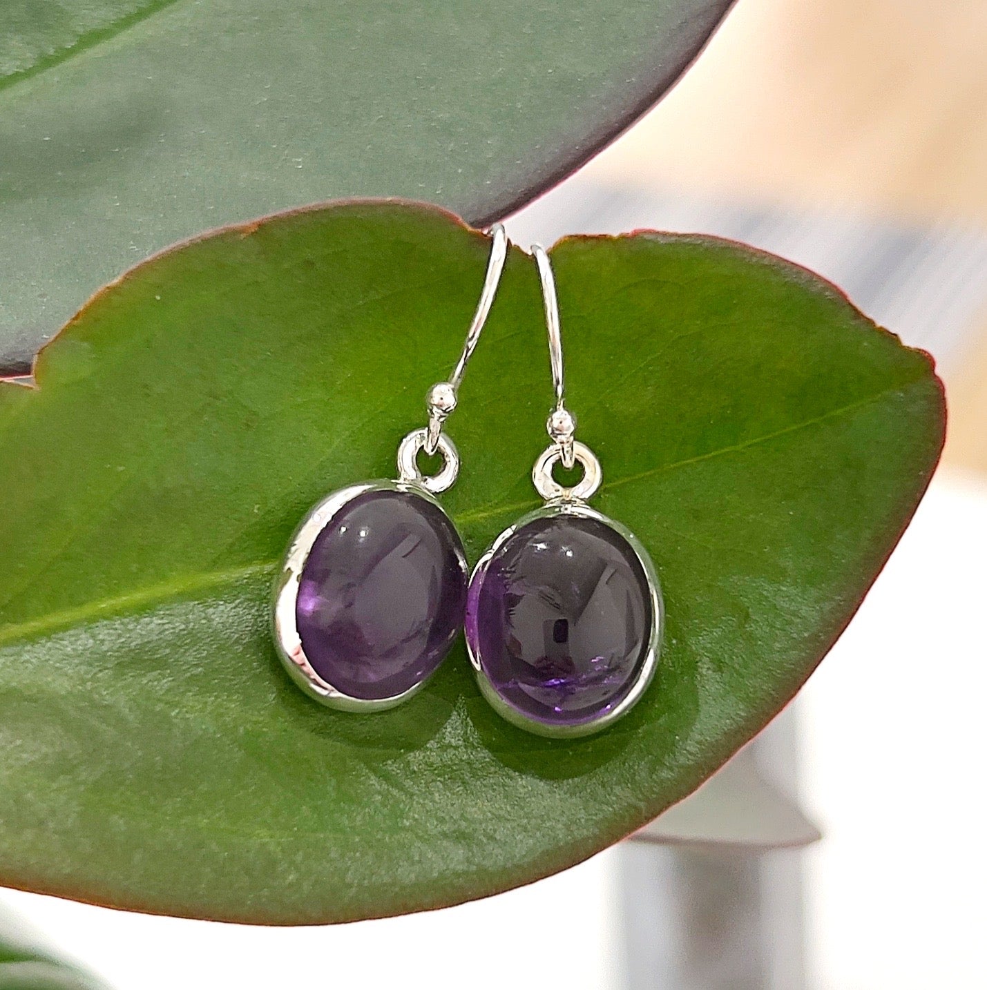 A pair of amethyst sterling silver drop earrings featuring oval-shaped, deep violet amethyst gemstones set in a smooth sterling silver bezel, displayed against a fresh green leaf background. The earrings have a simple hook design for a classic and elegant look. | Simply Esoteric