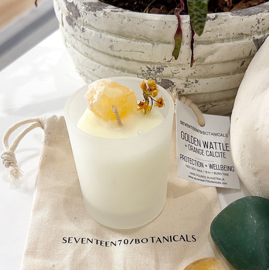 A soy wax candle from Seventeen70 Botanicals, infused with golden wattle and yellow calcite crystals, promoting protection and wellbeing. The candle sits beside a small bag and decorative stones, exuding an inviting ambiance. The label highlights its vegan-friendly attributes and a burn time of 18+ hours. | Simply Esoteric