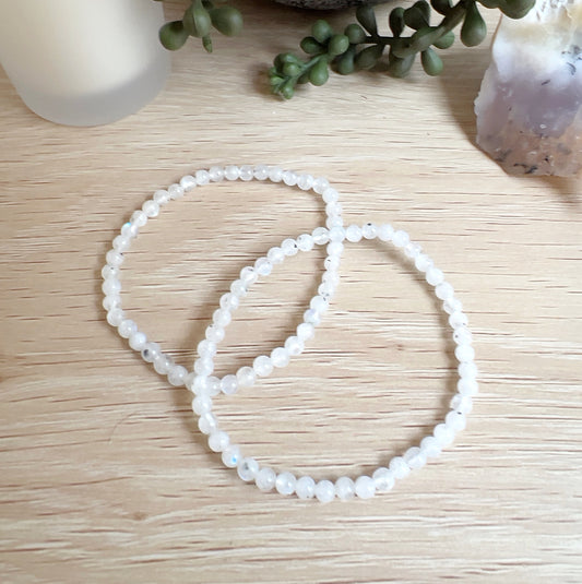 Two delicate moonstone crystal bead bracelets made with 4mm round beads, displayed on a wooden surface with a frosted candle, a leafy potted plant, and a piece of dendritic opal in the background. The beads are white with subtle flashes of blue, strung on stretchy cord. | Simply Esoteric