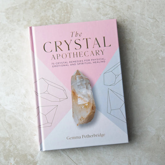 The image displays a book titled 'The Crystal Apothecary' by Gemma Petherbridge. The cover has a soft pink background with illustrations of crystal shapes and features a central image of a clear quartz point. The subtitle reads '75 Crystal Remedies for Physical, Emotional and Spiritual Healing', emphasizing the book's focus on holistic wellness through crystals. | Simply Esoteric