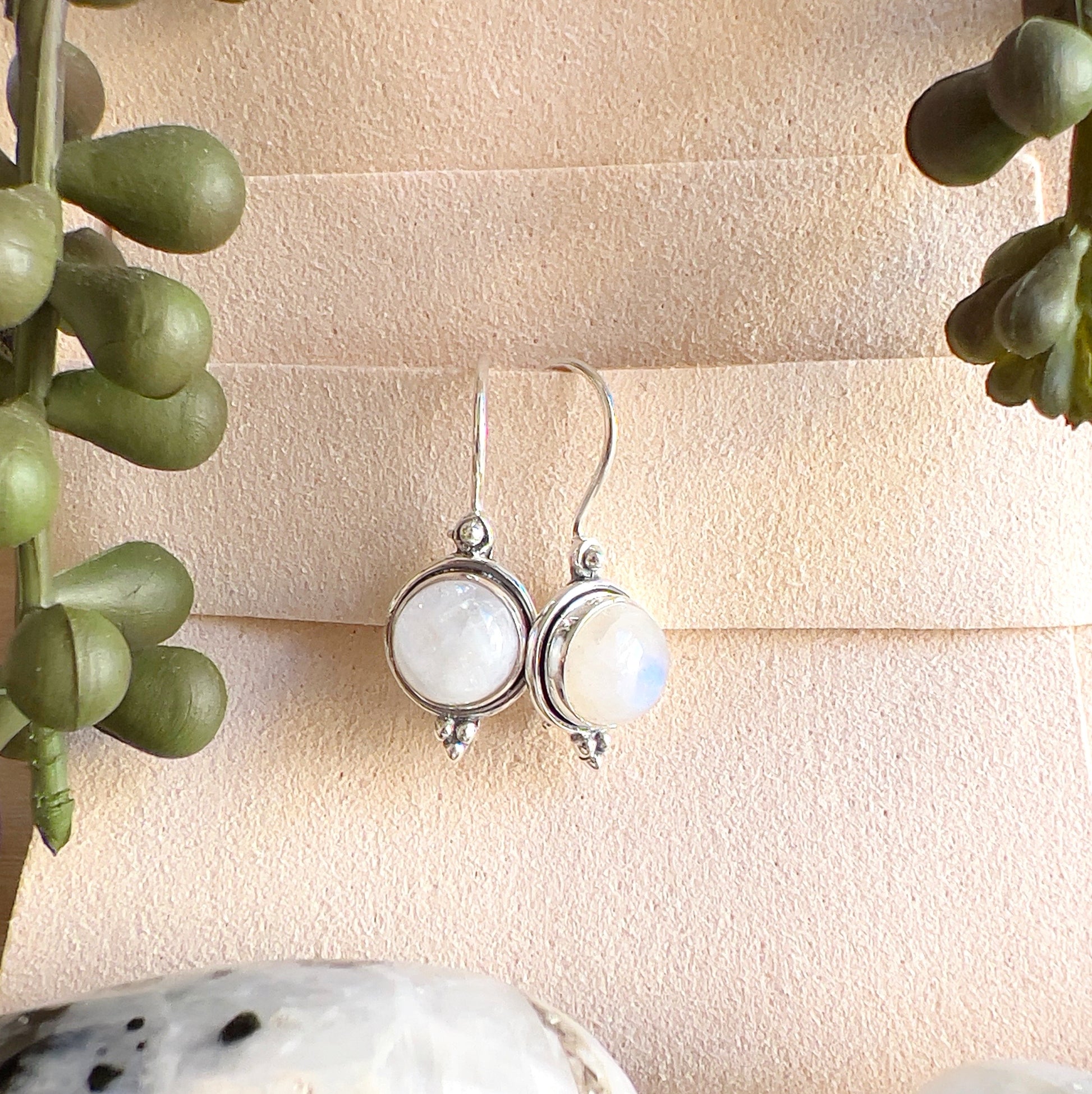 Sterling silver Moonstone earrings with round white stones showing soft flashes of blue, displayed on a light background with greenery accents. | Simply Esoteric