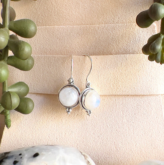 Sterling silver Moonstone earrings with round white stones showing soft flashes of blue, displayed on a light background with greenery accents. | Simply Esoteric