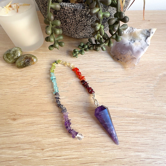 Amethyst pendulum with a faceted point and a chakra crystal chip chain, displayed on a light wooden surface alongside a candle, green tumbled stones, a potted plant, and a raw crystal cluster. | Simply Esoteric