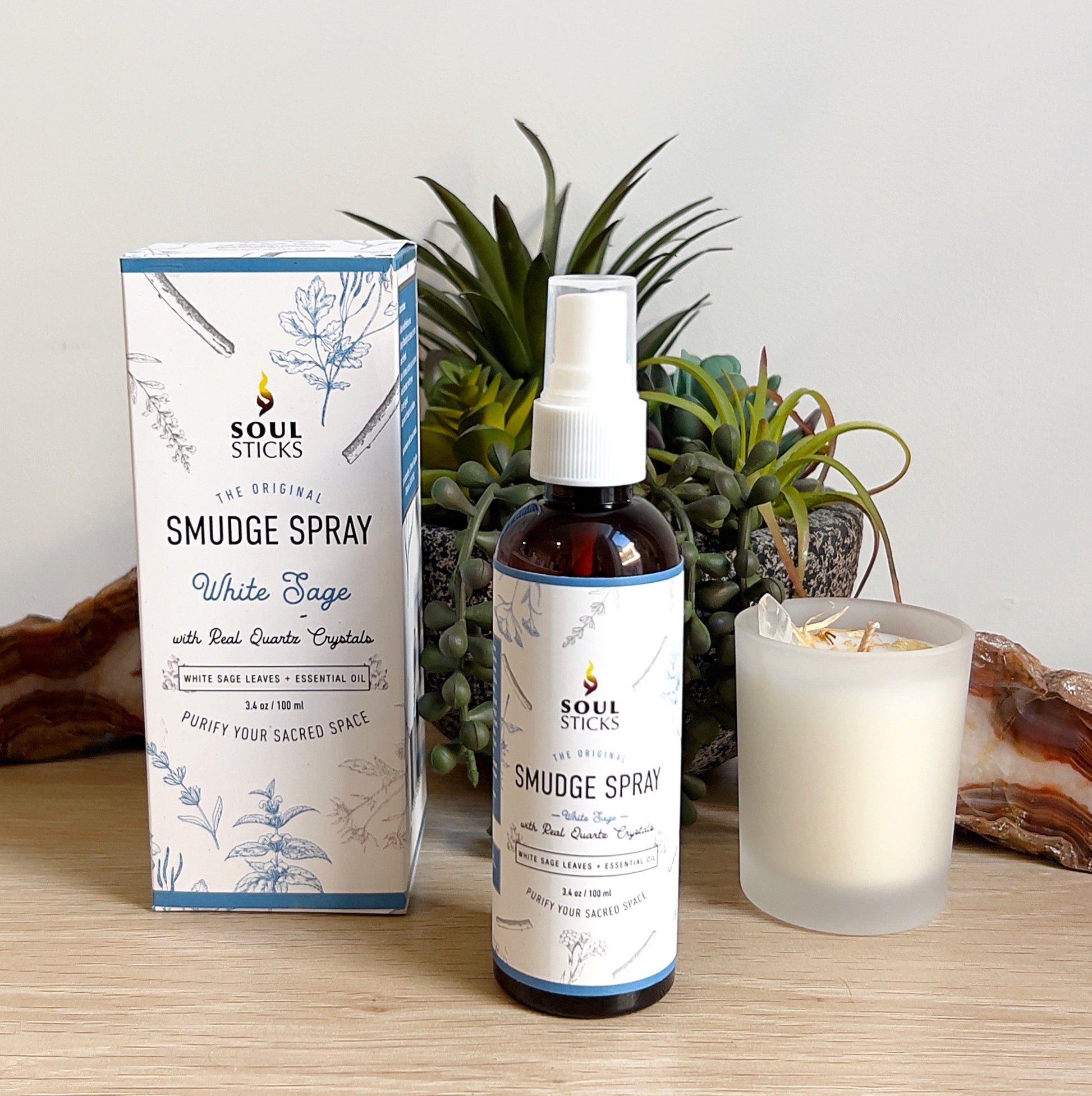 A 100ml bottle of Soul Sticks White Sage Smudge Spray with its box packaging, featuring white sage leaves and quartz crystals. The spray is displayed on a wooden surface beside a white candle, succulent plant, and polished agate slice, creating a clean and calming scene. | Simply Esoteric