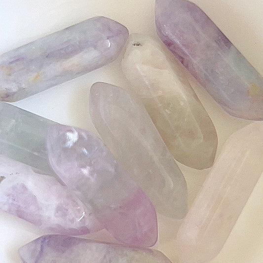 A collection of double-terminated rainbow fluorite points arranged loosely on a light background. The translucent crystals exhibit a gradient of soft purples, blues, and clears, blending together to create a gentle, calming spectrum of pastel hues. | Simply Esoteric