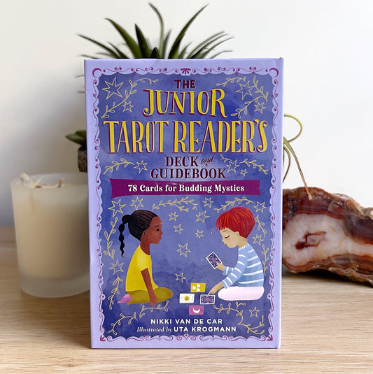 A colourful box of “The Junior Tarot Readers Deck and Guidebook” by Nikki Van De Car, featuring illustrated children reading tarot cards. Displayed on a wooden surface with a candle, succulent plant, and agate slice in the background, creating a magical and child-friendly setting. | Simply Esoteric