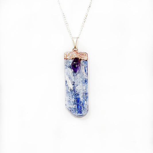 A striking pendant featuring a long, rectangular slab of polished blue kyanite topped with a small, deep purple amethyst crystal, set in a silver frame with intricate copper detailing, suspended from a silver chain. | Simply Esoteric