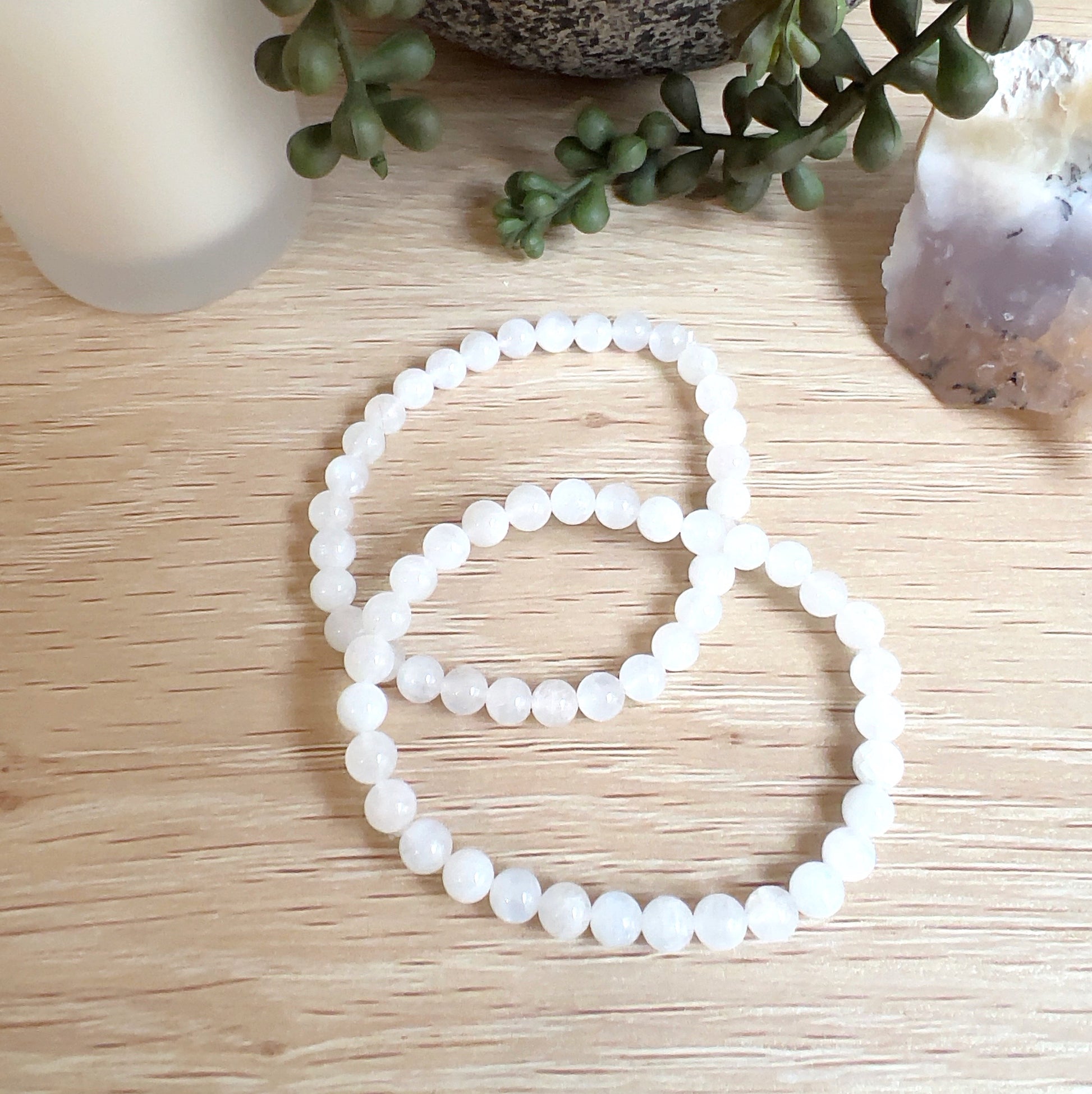 Two moonstone bead bracelets with 6mm round white beads on stretchy cord, displayed on a light wooden surface beside a candle, plant, and a piece of dendritic opal. | Simply Esoteric