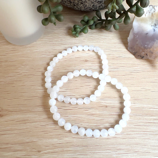 Two moonstone bead bracelets with 6mm round white beads on stretchy cord, displayed on a light wooden surface beside a candle, plant, and a piece of dendritic opal. | Simply Esoteric