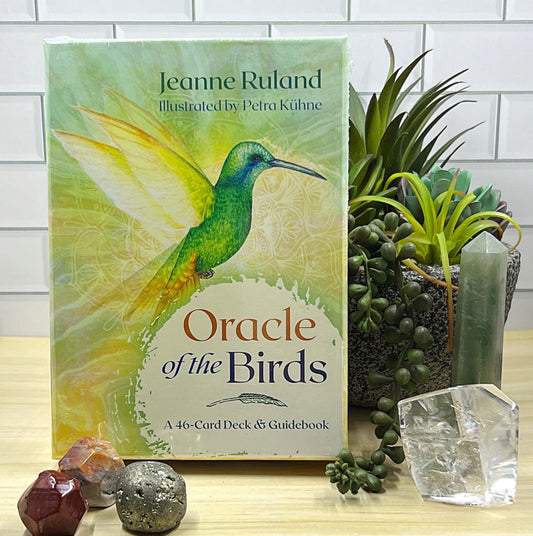 Oracle of the Birds card deck by Jeanne Ruland displayed on a tabletop surrounded by crystals and succulents. The box features vibrant artwork of a green and yellow hummingbird in flight, with the title 'Oracle of the Birds' and subtitle 'A 46-Card Deck & Guidebook' visible on the front. | Simply Esoteric