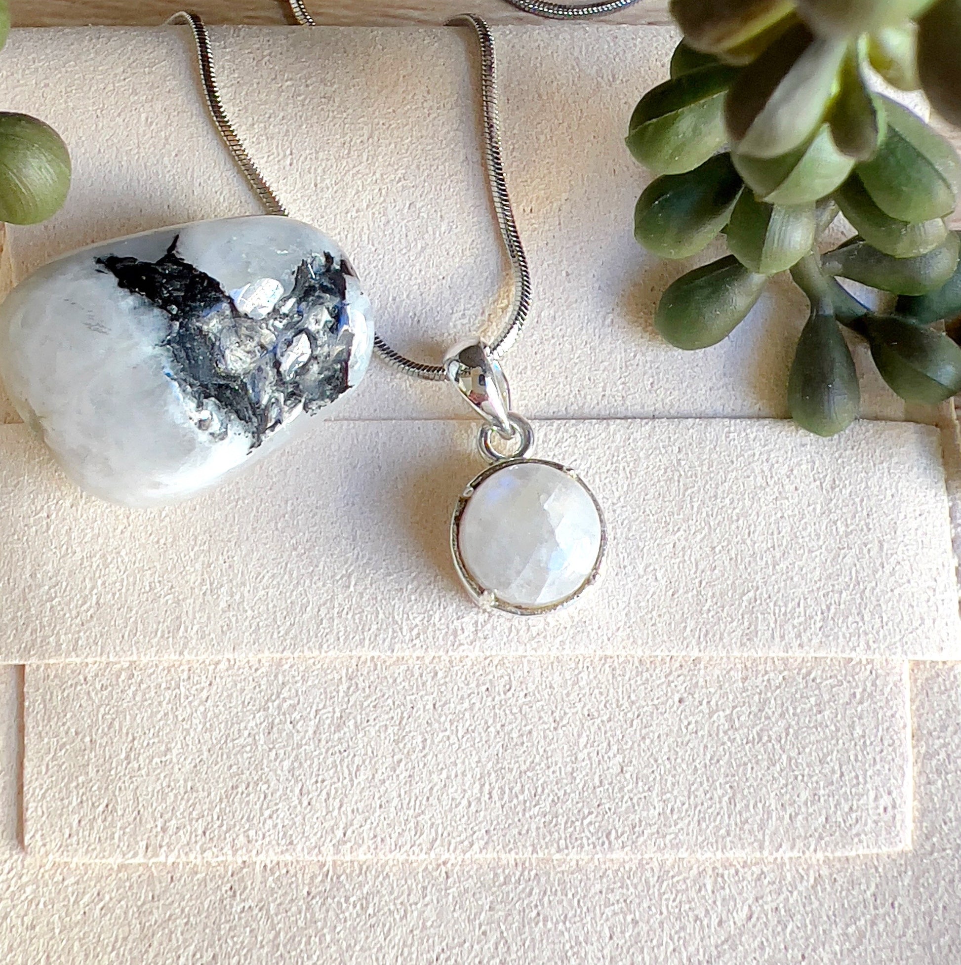 Sterling silver Moonstone pendant with a luminous round stone, displayed on a silver chain alongside polished Moonstone crystals on a light surface. | Simply Esoteric